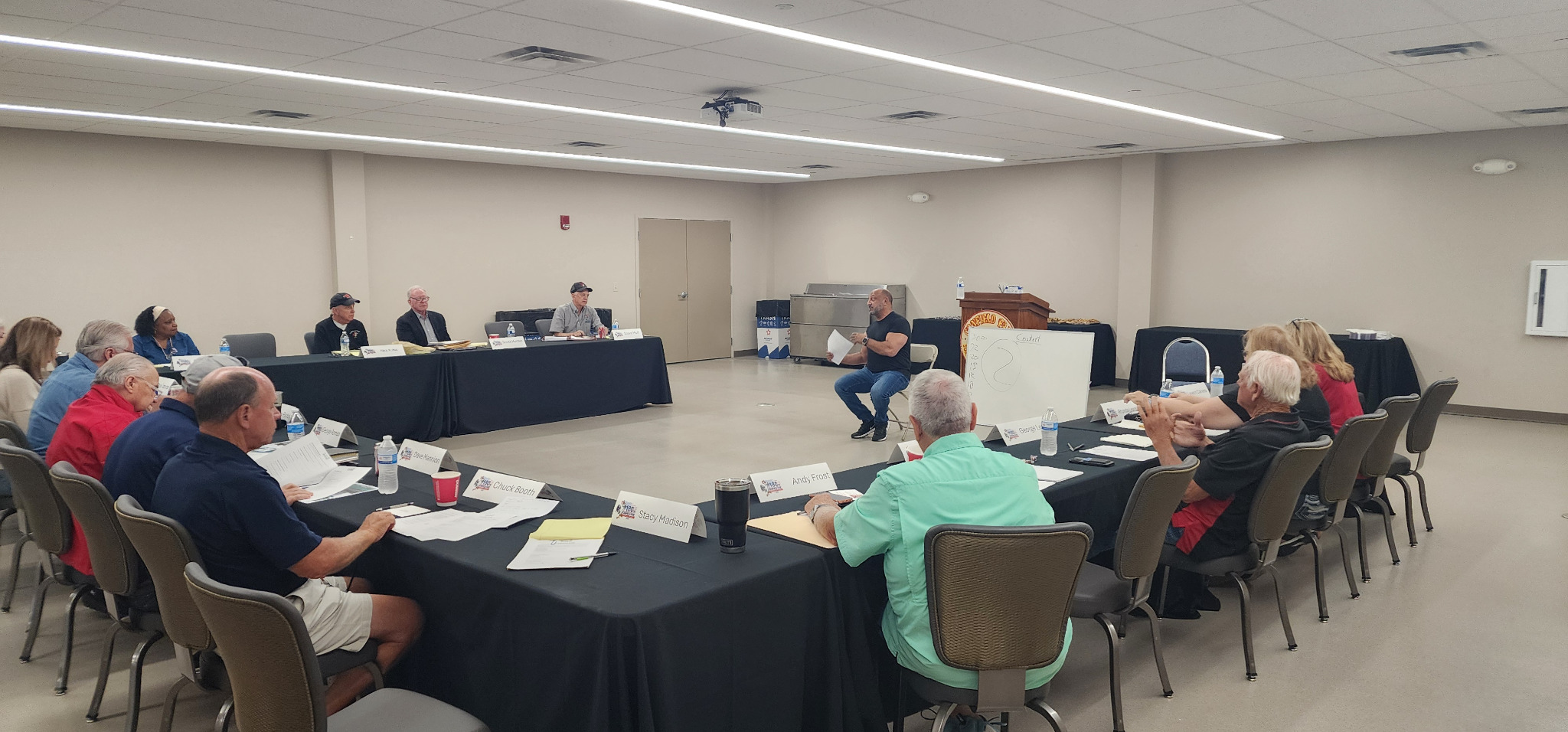 Participants discuss workshop materials around U-shaped tables while a facilitator leads the session.
